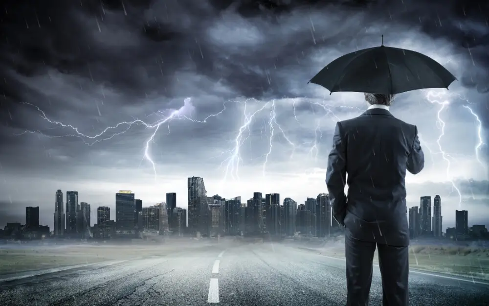 Businessman with an umbrella standing on a road facing a city skyline under a dark severe thunderstorm with lightning bolts, representing corporate weather risk.